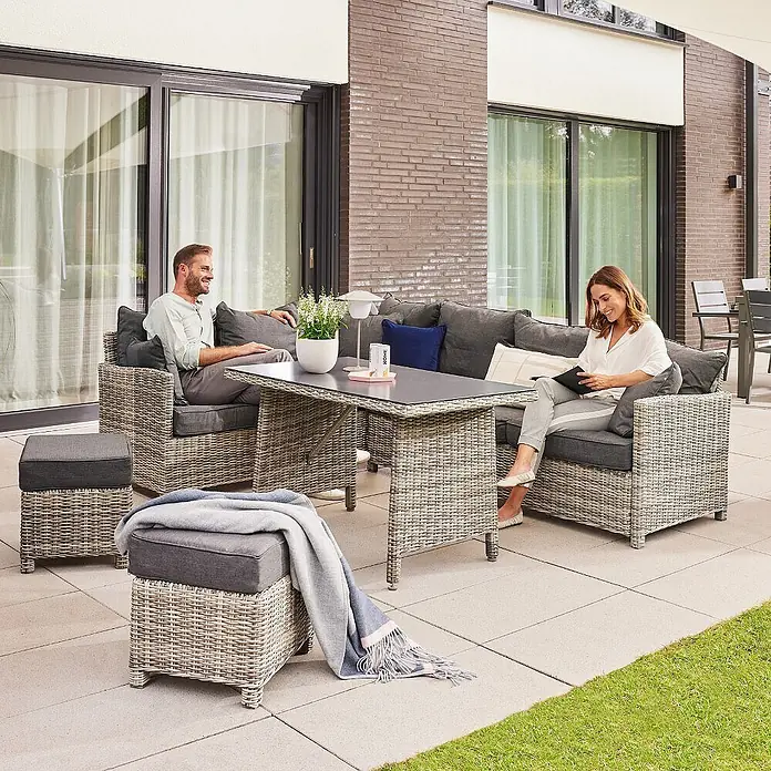 Home Deluxe Polyrattan Sitzgruppe BAHIA Mit Regenabdeckung -  - Graues Rattan-Gartenlounge-Set mit dunklen Kissen und einem Tisch, von zwei Personen auf einer Terrasse genutzt.