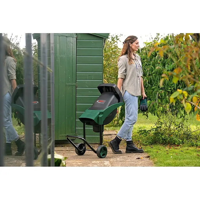 Bosch Elektro-Messerhäcksler Axt Rapid 2000 - Max. Aststärke: 3,5 cm, 2.000 W - Grün-schwarzer Bosch AXT Rapid Gartenhäcksler auf Rädern, einsatzbereit. Eine Frau hält Gartenhandschuhe bereit.