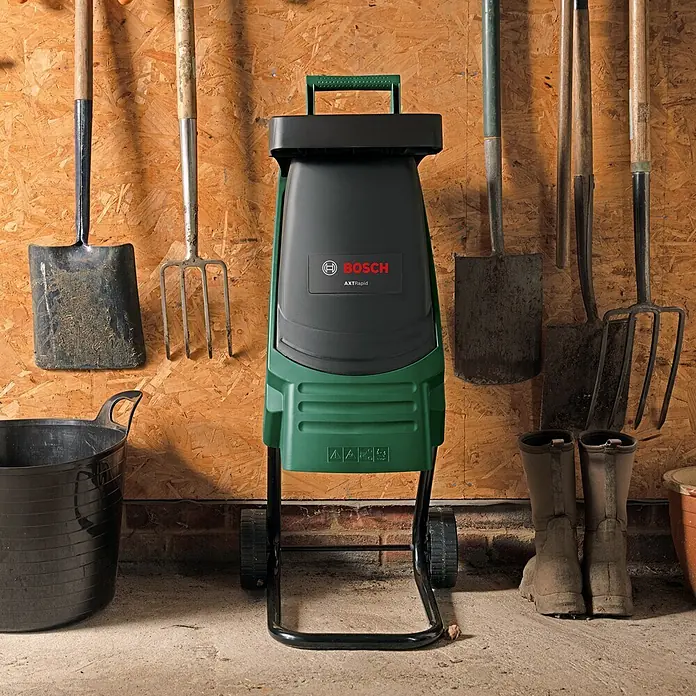 Bosch Elektro-Messerhäcksler Axt Rapid 2000 - Max. Aststärke: 3,5 cm, 2.000 W - Grüner und schwarzer Bosch AXT Rapid Gartenhäcksler mit Rädern, in einem Schuppen zwischen Gartengeräten und Stiefeln.