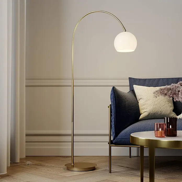 Lindby Stehleuchte Cynthia - Höhe: 140 cm, messing / gold, E27 - Gebogene Messing-Stehlampe mit weißem Kugeldiffusor beleuchtet eine gemütliche Wohnzimmerecke neben einem blauen Sofa.