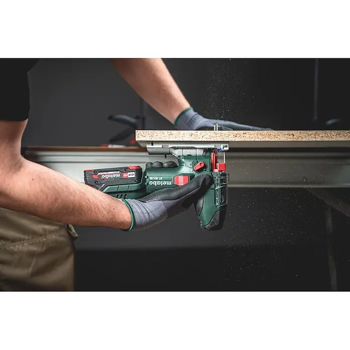 Metabo CAS 18V Akku-Pendelhubstichsäge ST 18 L 90 - 18 V, Anzahl Akkus: Ohne Akku, 750 /min - 3.000 /min - Grüne und schwarze Metabo Akku-Stichsäge schneidet helle Spanplatte, von behandschuhter Hand gehalten. Sägemehl fliegt.