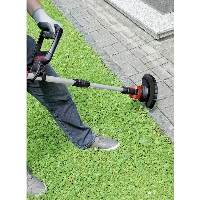 Einhell PXC 18V Recortabordes de batería GE-CT 18 Li-Solo - 18 V, Iones de litio, Sin batería - Un hombre usa un cortacésped eléctrico para cortar el césped a lo largo de un camino pavimentado. El cortacésped es rojo y negro con un eje gris.