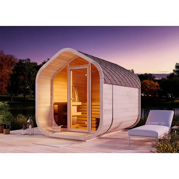 FinnTherm Fasssauna ScandiCube Wave Comfort - Holz - Helle Holz-Außensauna mit dunklem Dach auf einer Terrasse, abends von innen beleuchtet, neben einem Liegestuhl.