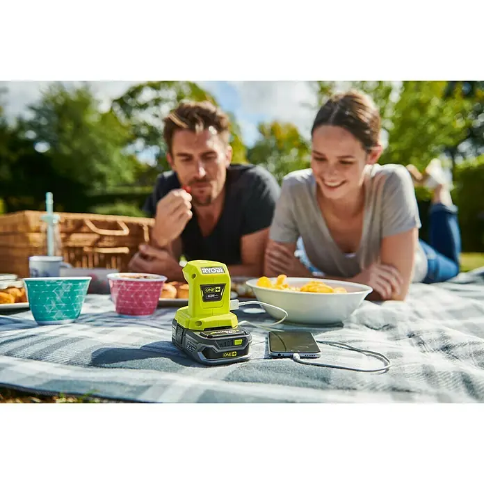Ryobi ONE+ USB-Adapter - 18 V, Ohne Akku - Ryobi ONE+ Akku-Ladegerät und Smartphone auf einer Picknickdecke, mit Paar im Hintergrund. Praktisch für unterwegs!