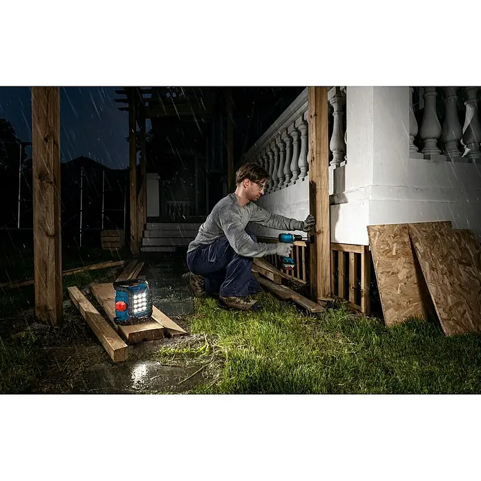 Bosch Professional AMPShare 18V LED-Akkustrahler PRO GLI18V-1500 - 18 V, Ohne Akku, 1.500 lm - Mann befestigt Holz mit blaugrünem Akku-Bohrschrauber in regnerischer Nacht, beleuchtet von Arbeitsleuchte.