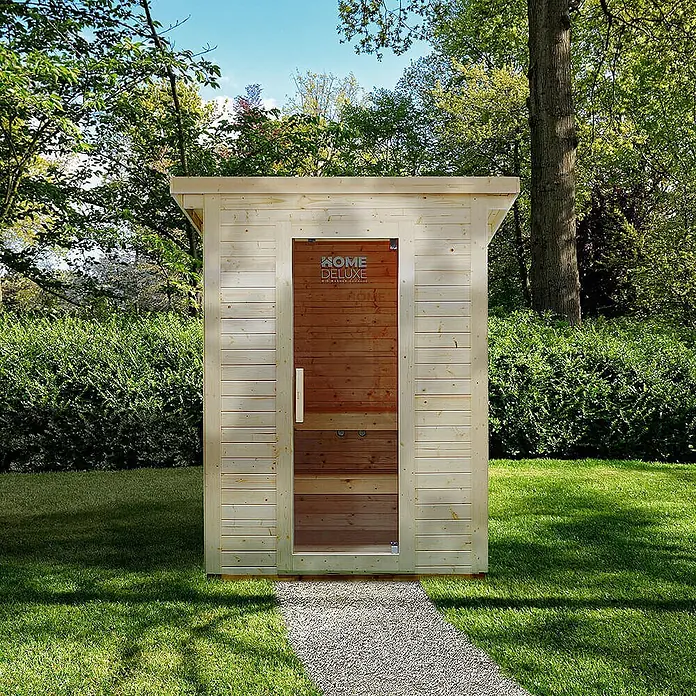 Home Deluxe Fasssauna TALO M Größe: M - Elektroofen, Holz - Hellholz-Außensauna mit Glastür und dunkler Holzverkleidung, auf einem Kiesweg im grünen Garten.