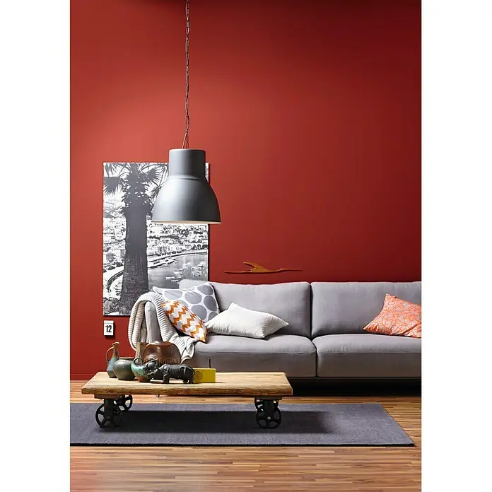 SCHÖNER WOHNEN-Farbe Wandfarbe Trendfarbe - Amarena, Matt, 2,5 l, Konservierungsmittelfrei - Wohnzimmer mit einer grauen Industrielampe, einem grauen Sofa und einem Holztisch mit Rädern. Die rote Wand sorgt für einen warmen Akzent.