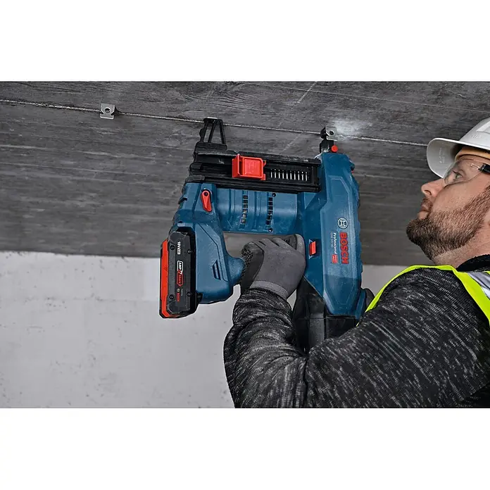 Bosch Professional Deckenhaken PRO FMH - 100 Stk. - Person verwendet ein blau-rotes Bosch Professional 18V Nagelsystem, um eine Metallklammer an einer Betondecke zu befestigen.