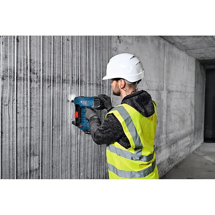 Bosch Professional Unterlegscheibe PRO FMW-25 - Metall, 1.000 Stk. - Ein Arbeiter mit Schutzhelm und Warnweste befestigt mit einem blauen Bosch-Nagelgerät etwas an einer gerippten Metallwand.