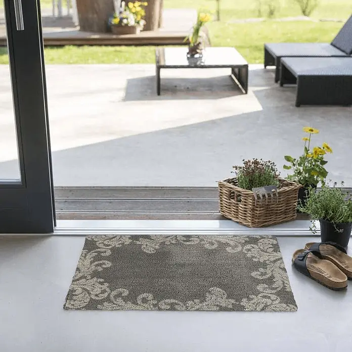 Akzente Fußmatte Feel - Mehrfarbig, Motiv / Muster|Floral - Eine graue Fußmatte mit hellem, verziertem Muster, an einer offenen Terrassentür platziert, mit Schuhen und Pflanzen daneben.