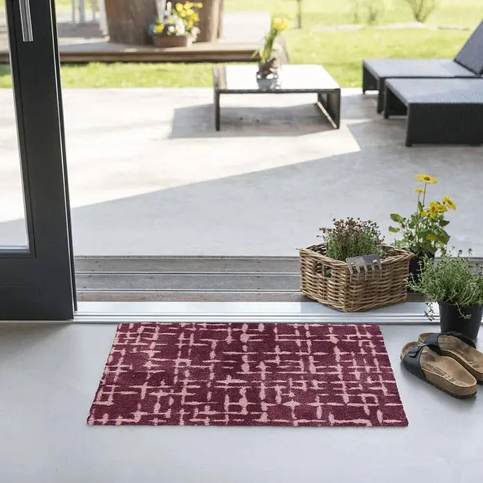 Akzente Fußmatte Feel - 70 x 50 cm, Mehrfarbig, Motiv / Muster - Dunkelrote Fußmatte mit abstraktem Muster am Eingang einer Schiebetür, umgeben von Pflanzen und Sandalen.