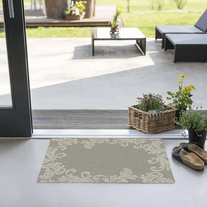Akzente Fußmatte Feel - Mehrfarbig, Motiv / Muster|Floral - Eine hellgraue Fußmatte mit einem eleganten, hellen Zierrandmuster, platziert an einem Glaseingang zu einer Terrasse.
