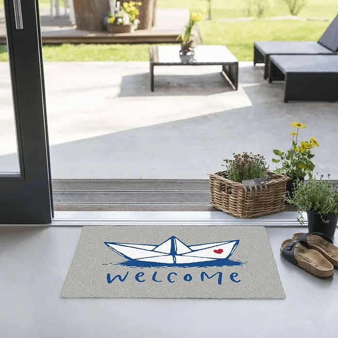 Akzente Fußmatte Feel - Mehrfarbig, Motiv / Muster|Schriftzug - Graue Fußmatte mit blauer Papierboot-Illustration mit rotem Herz und „welcome“-Schriftzug, an einem Terrasseneingang platziert.