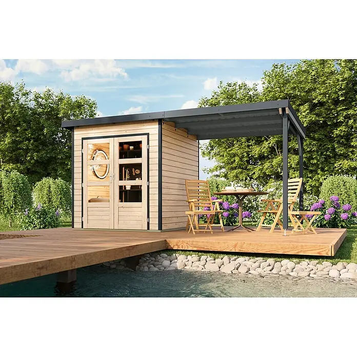 Karibu Gartenhaus Pluto 1 A - Außenmaß inkl. Dachüberstand (B x T): 447 x 228 cm, Holz, Naturbelassen/Anthrazit - Gartenhaus aus hellem Holz mit dunkelgrauer integrierter Pergola auf einer Holzterrasse am Wasser. Gartenmöbel darunter.