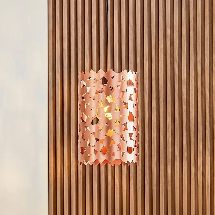 Lucande Pendelleuchte Aeloria - kupfer, Kupfer, E27 - Kupfer-Pendelleuchte mit perforiertem zylindrischem Schirm, vor einer Holzlamellenwand hängend.