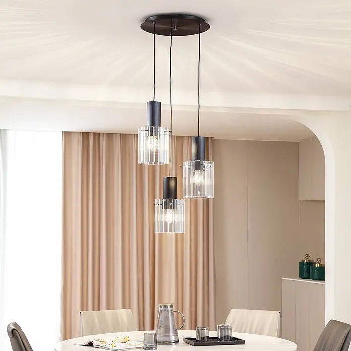 Lucande Pendelleuchte in transparent, schwarz, Hängelampe -  - Drei moderne Pendelleuchten mit dunklen zylindrischen Oberteilen und klaren gerippten Glasschirmen hängen über einem Esstisch.