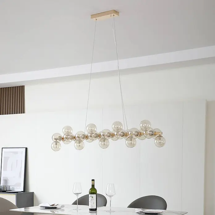 Lucande Pendelleuchte Naelen - L x B x H: 125 x 24,5 x 20 cm, creme / amber, gold, cognac, G9 - Lange lineare Pendelleuchte mit bernsteinfarbenen Glaskugeln und goldfarbenem Rahmen über einem Esstisch.