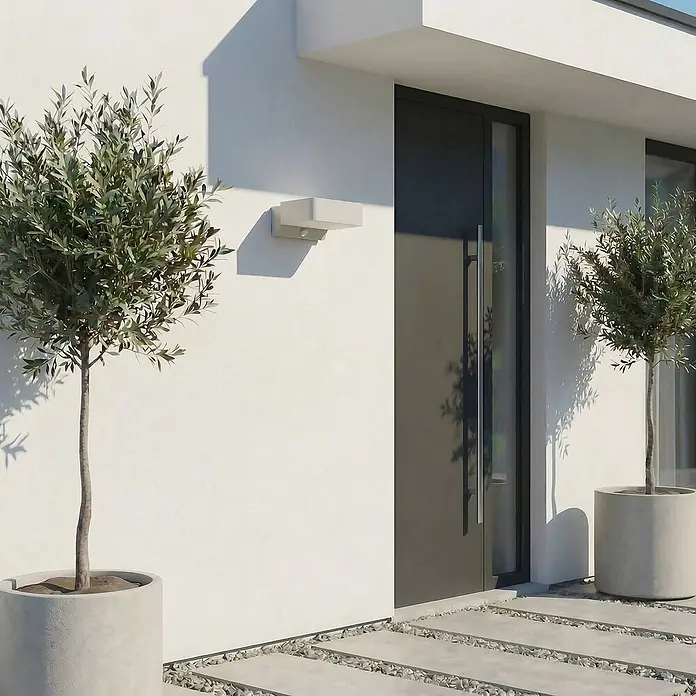 Lindby Außenwandleuchte Fando - 14,7 x 22,5 x 8,8 cm, alu / grau / zink, IP44 - Moderne, hellfarbene Außenwandleuchte an einer weißen Fassade neben einer dunklen Tür zur Eingangsbeleuchtung.