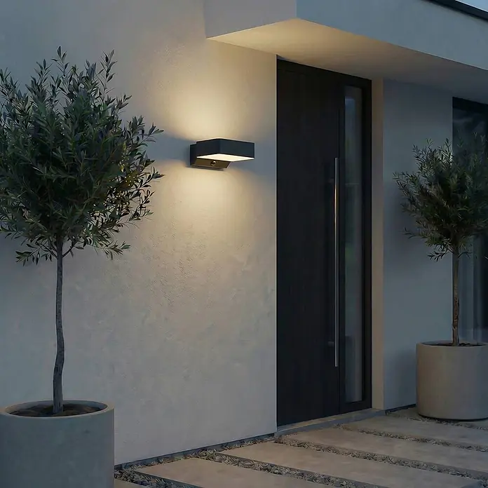 Lindby Außenwandleuchte Fando - 14,7 x 22,5 x 8,8 cm, alu / grau / zink, IP44 - Dunkle rechteckige Außenwandleuchte beleuchtet einen Gebäudeeingang mit warmem Licht.