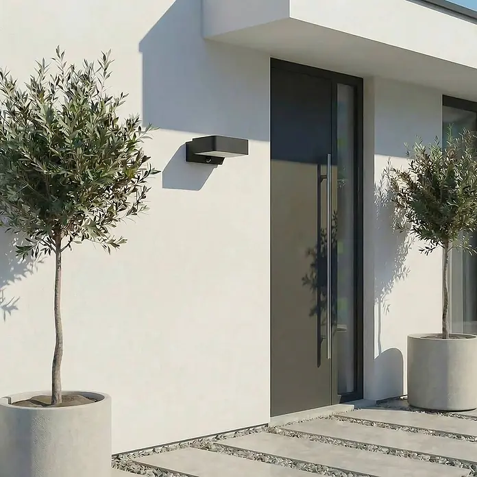 Lindby Außenwandleuchte Fando - 14,7 x 22,5 x 8,8 cm, alu / grau / zink, IP44 - Schwarze, rechteckige Außenwandleuchte an einer modernen Hausfassade, beleuchtet den Eingang neben einem Olivenbaum.