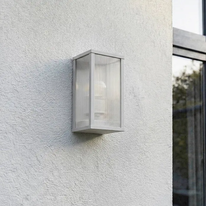Lindby Außenwandleuchte Peldar - 13,5 x 14 x 26 cm, alu / grau / zink, IP44 - Rechteckige, hellgraue Außenwandleuchte mit geriffelten Klarglaspaneelen, montiert an einer strukturierten Außenwand.