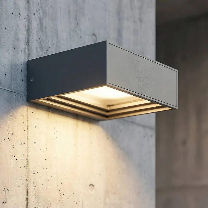 Lucande Außenwandleuchte Ashkan - alu / grau / zink, IP65 - Dunkelgraue, rechteckige Außenwandleuchte an Betonwand, wirft warmes Licht nach unten.