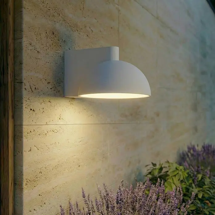 Lucande Außenwandleuchte Velira - 20 x 16 x 11 cm, alu / grau / zink, alu / grau / zink, IP54 - Weiße, gewölbte Außenwandleuchte beleuchtet eine strukturierte Steinwand und Lavendelpflanzen.