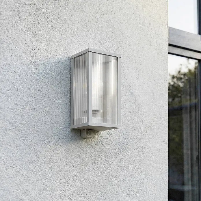 Lindby Außenwandleuchte Peldar - alu / grau / zink, IP44 - Graue rechteckige Außenwandleuchte mit Riffelglas und Bewegungsmelder, an Gebäudefassade montiert.