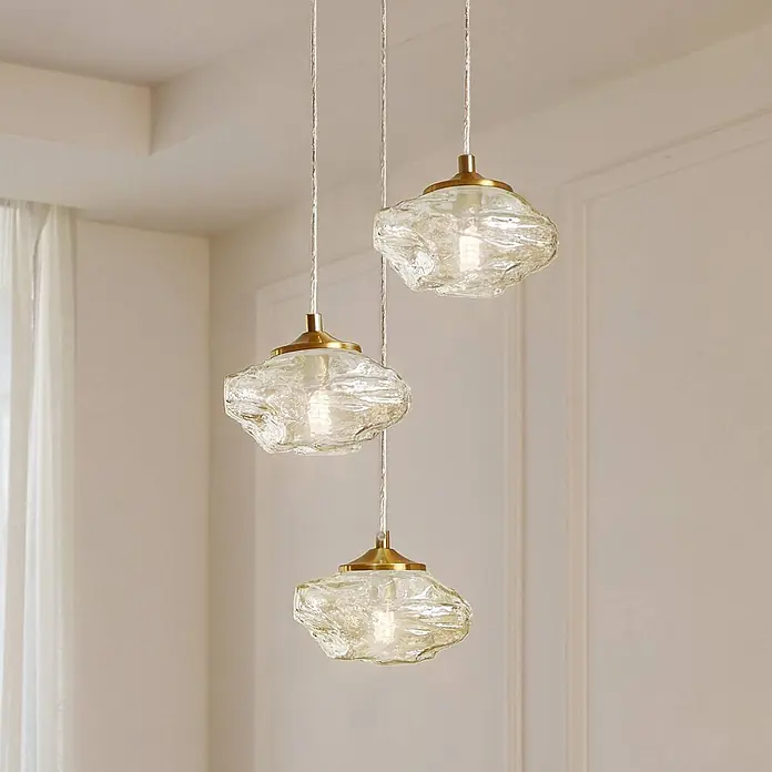 Lindby Pendelleuchte Piatra - L x B x H: 25 x 25 x 150 cm, creme / amber, amber, messing, G9 - Drei Pendelleuchten aus strukturiertem Klarglas mit Messingbeschlägen hängen in einem modernen Raum von der Decke.