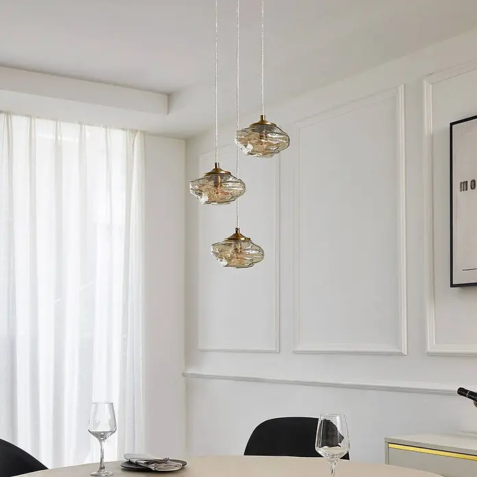 Lindby Pendelleuchte Piatra - L x B x H: 25 x 25 x 150 cm, creme / amber, amber, messing, G9 - Drei moderne Pendelleuchten mit strukturierten bernsteinfarbenen Glasschirmen und Messingakzenten hängen über einem Esstisch.