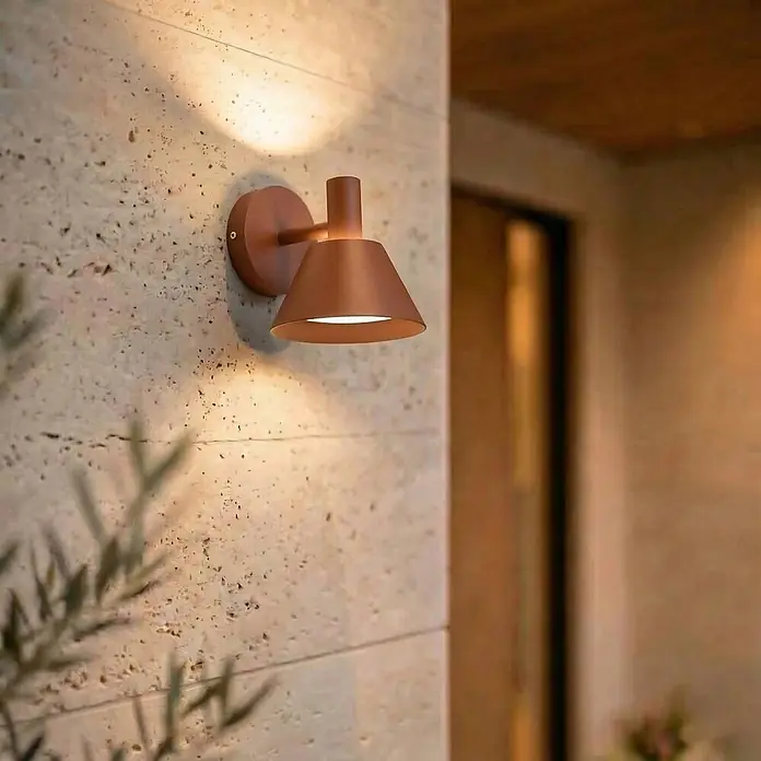 Lucande Außenwandleuchte Tabena - braun / rost, braun / rost, IP54 - Kupferfarbene, kegelförmige Wandleuchte beleuchtet eine strukturierte Steinwand mit warmem Licht.