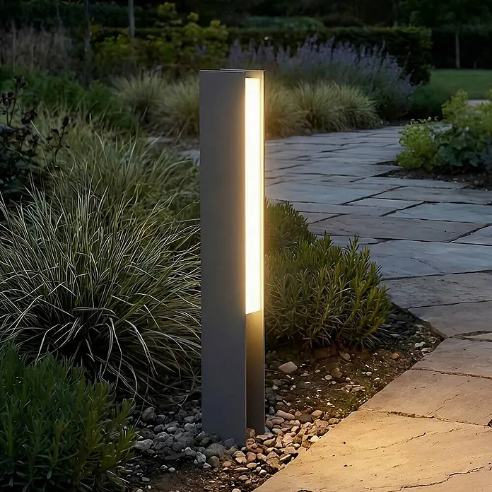 Lucande LED Sockelleuchte in schwarz, Wegeleuchten außen -  - Graue Außenpollerleuchte beleuchtet einen Gartenweg inmitten von Pflanzen.