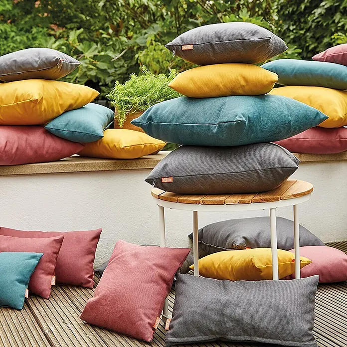 Siena Garden Kissen PRIME - 45 x 45 cm, Anthrazit, Quadratisch - Gestapelte und verstreute Outdoor-Kissen in Grau, Gelb, Petrol und Rosa, auf einer Terrasse.