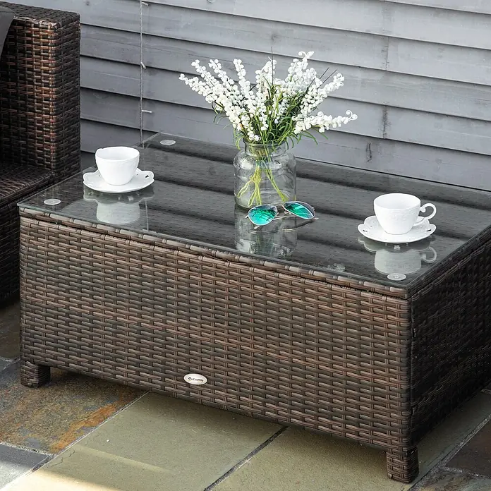 Outsunny Garten-Loungetisch - Metall, PE-Rattan, gehärtetes Glas, Braun, L x B x H: 85 x 50 x 39 cm - Brauner Korb-Couchtisch für den Außenbereich mit Glasplatte, auf einer Terrasse mit Tassen und Blumen.