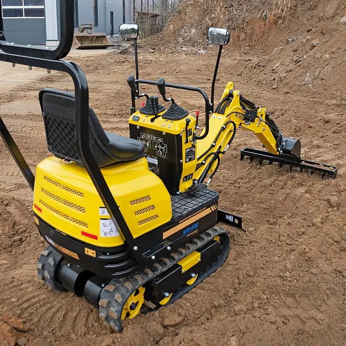 Bamato Minibagger MIB-600 - 6.300 W, Benzin, Auslegerschwenkbereich: 70 ° - Gelber und schwarzer Raupen-Minibagger mit Rechenaufsatz auf Erdboden.