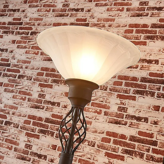 Lucande LED-Deckenfluter Dunja - braun / rost - Leuchtende Stehlampe mit mattem Glasschirm und dunklem Metall-Spiralgestell vor einer Ziegelwand.