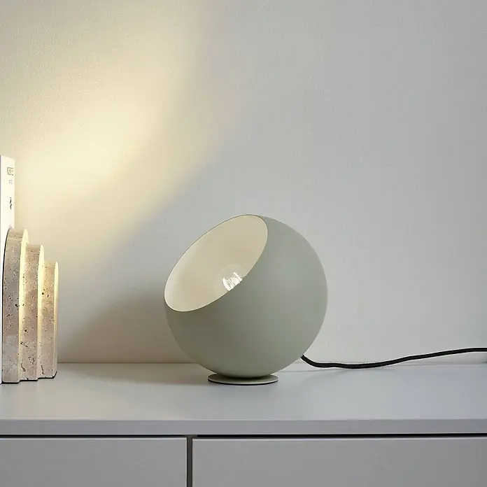 Lindby Tischleuchte Solomia - L x B x H: 20 x 20 x 20 cm, grün, E27 - Salbeigrüne kugelförmige Tischleuchte mit sichtbarer Glühbirne und schwarzem Kabel, die eine Oberfläche neben Deko-Objekten beleuchtet.