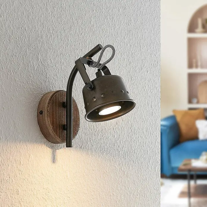 Lindby Wandleuchte Rubinjo - schwarz, GU10 - Industrieller Wandstrahler mit dunklem Metallgehäuse, Holzsockel und geflochtenem Kabel, strahlt warmes Licht aus.