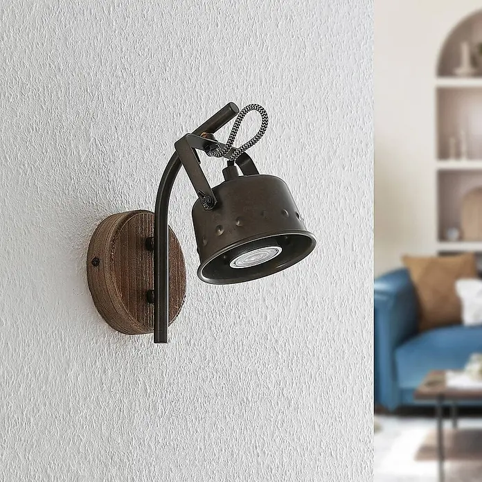 Lindby Wandleuchte Rubinjo - schwarz, GU10 - Wandleuchte im Industriestil mit dunklem Metallstrahler, Holzsockel und geflochtenem Kabel, an einer strukturierten Wand montiert.