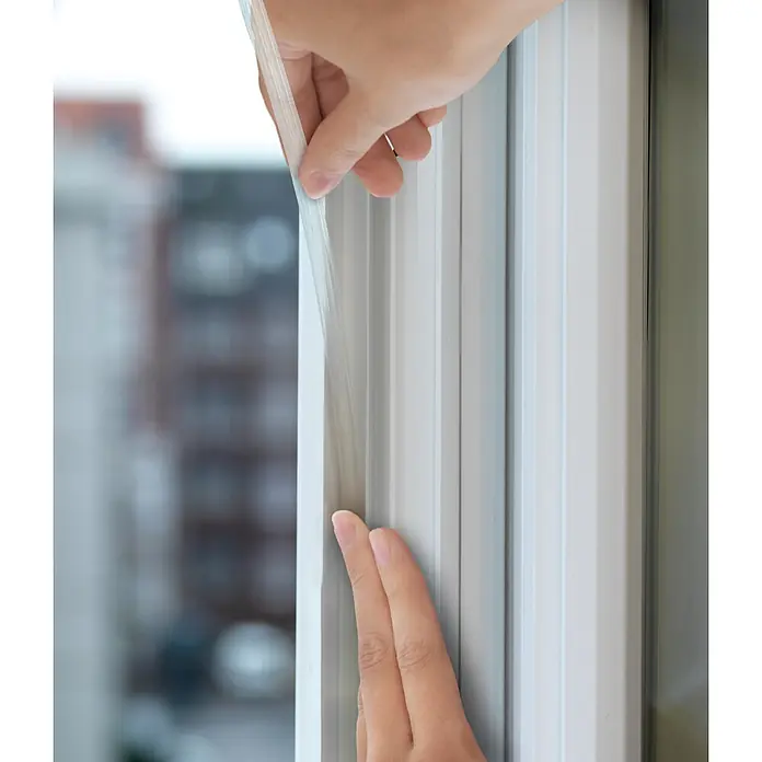 Tesa INSULATION Silikondichtung - Transparent, 6 m x 9 mm x 7 mm, Geeignet für: Spaltenbreiten 1 - 7 mm - Hände bringen einen transparenten, flexiblen Dichtungsstreifen an einem weißen Fensterrahmen an.