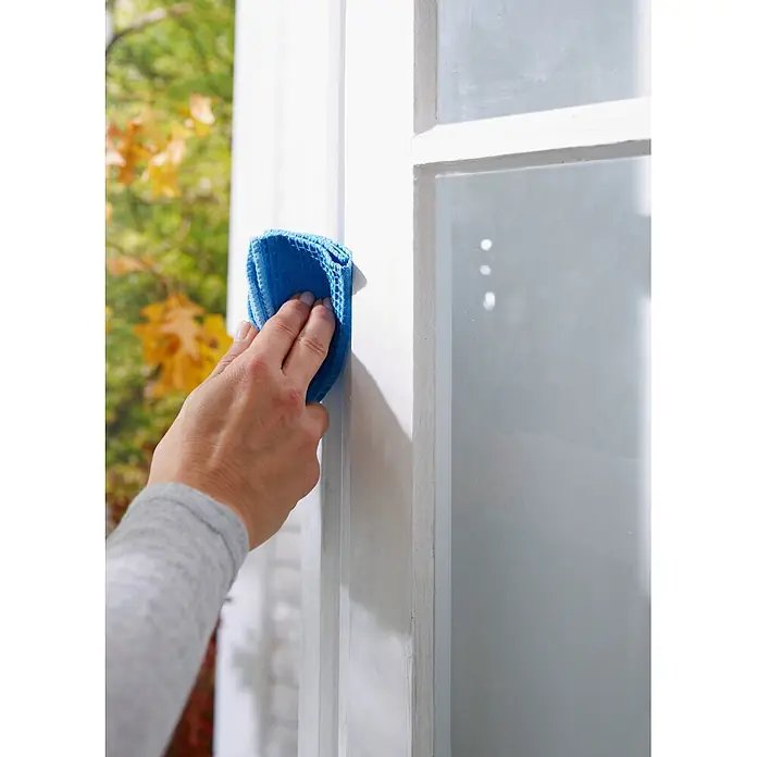 Tesa INSULATION Dichtungsband GUMMI L - Weiß, 10 m x 9 mm x 5,5 mm, Geeignet für: Spaltenbreiten 2 - 5 mm - Hand reinigt einen weißen Fensterrahmen mit einem blauen Mikrofasertuch.