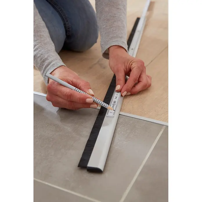 Tesa INSULATION Türdichtschiene für unebene Böden - Braun, Geeignet für: Spaltenbreiten bis 15 mm - Person markiert Bodenbelag mit grauem Führungsschiene und schwarzem Bürstenrand mit Bleistift.