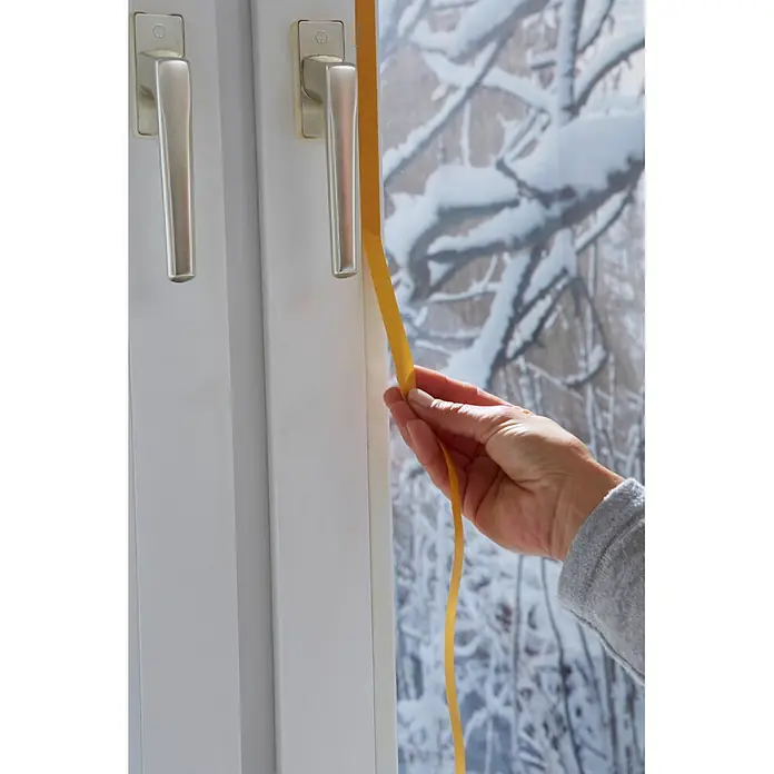 Tesa INSULATION Fensterisolierfolie - 4 x 1,5 m, Farblos, Transparent - Eine Hand bringt braunes Dichtungsband an einem weißen Fensterrahmen an, um gegen die verschneite Winteraussicht zu isolieren.