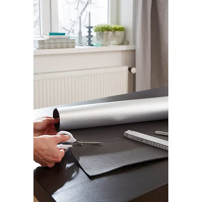 Tesa INSULATION Reflektorfolie für Heizkörper - Silber, 100 x 70 cm, Heizkörpernische - Person schneidet eine silberne, texturierte Isolier- oder Schutzmatte mit Schere auf einem dunklen Tisch, Lineal liegt daneben.