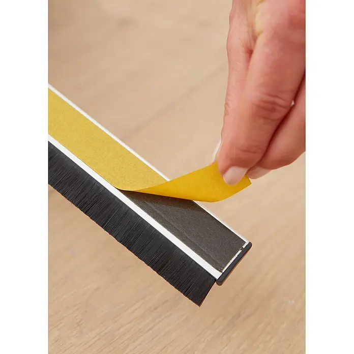 Tesa INSULATION Türdichtschiene für unebene Böden - Weiß, Geeignet für: Spaltenbreiten bis 15 mm - Hand zieht gelbe Schutzfolie von schwarzer Bürstendichtung mit Klebestreifen, bereit zur Montage auf Holzfußboden.