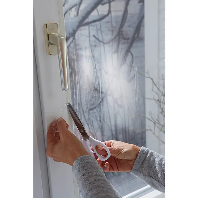 Tesa INSULATION Fensterisolierfolie - 4 x 1,5 m, Farblos, Transparent - Hände schneiden transparente Fensterisolierfolie mit weißer Schere am Fensterrahmen.