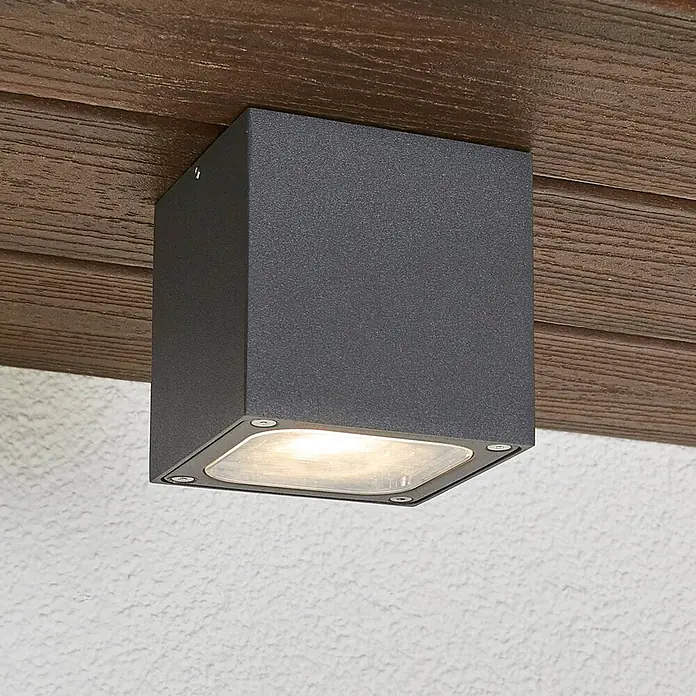 Lucande Außenwand- & Deckenleuchte Tanea - alu / grau / zink, alu / grau / zink, IP54 - Dunkelgraue, würfelförmige Außenleuchte an Holzbalken, wirft warmes Licht auf eine weiße Wand.