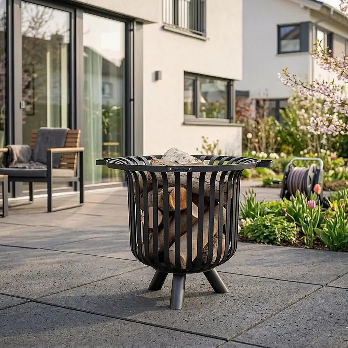 Bene on Fire Holzkohlegrill Tennessee - Schwarz - Schwarze Feuerschale aus Metall, gefüllt mit Holzscheiten, auf einer Terrasse vor Haus und Garten.