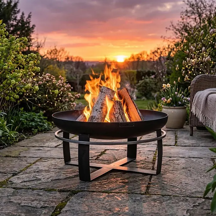 Bene on Fire Holzkohlegrill Maryland - Schwarz - Schwarze Metall-Feuerschale mit brennenden Holzscheiten auf einer Steinterrasse im Garten bei Sonnenuntergang.