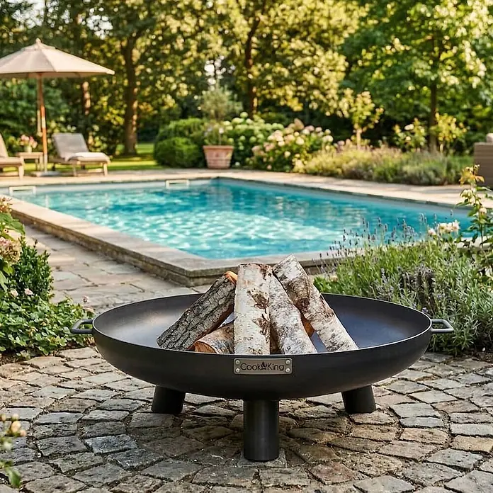Bene on Fire Holzkohlegrill Louisiana - schwarz - Schwarze Metall-Feuerschale mit Holzscheiten auf einer Steinterrasse neben einem Pool.